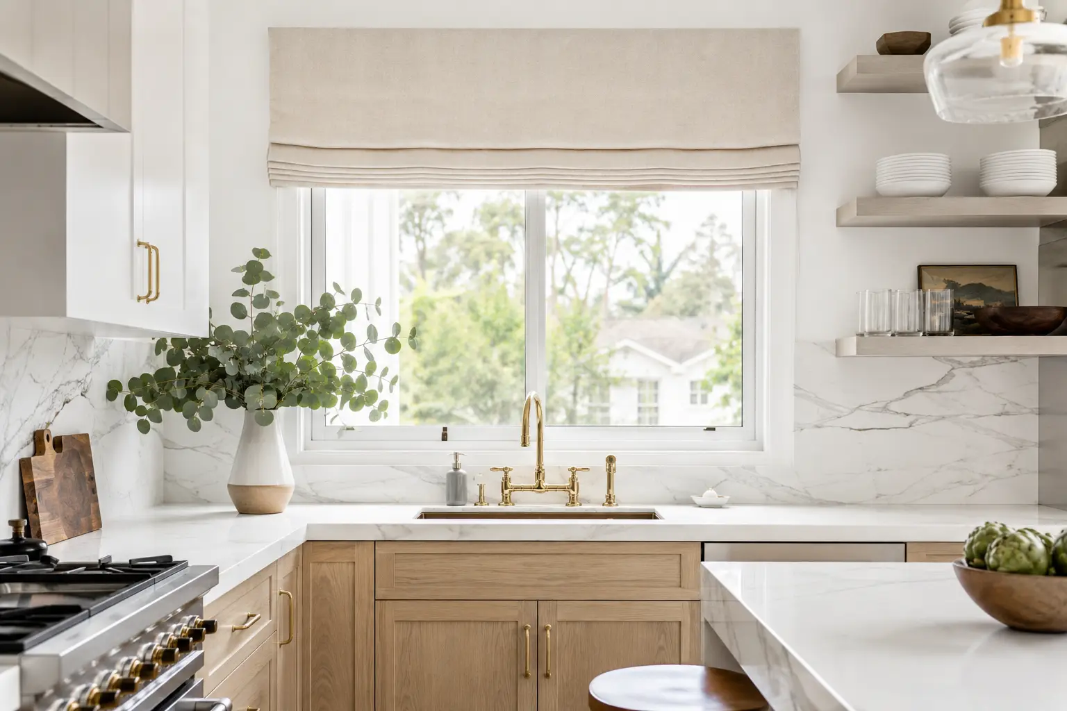 collaborating with a drapery workroom for custom window treatments. Bright modern farmhouse kitchen featuring a custom linen Roman shade over a window, a luxury gold faucet, marble countertops, and light oak cabinets.